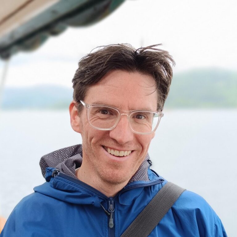 Picture of a man stood in front of a lake and smiling at the camera