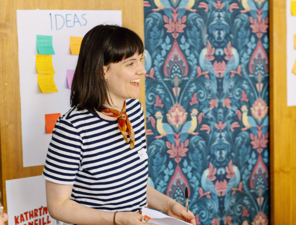 Woman stood in front of a piece of paper labelled 'Ideas' covered in sticky notes with writing on