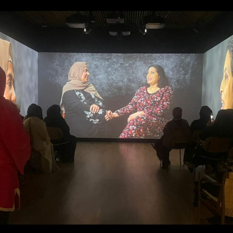 A small room in a museum with audience members watching a film that is being displayed on three of the four walls. The film depicts South Asian women talking to each other.